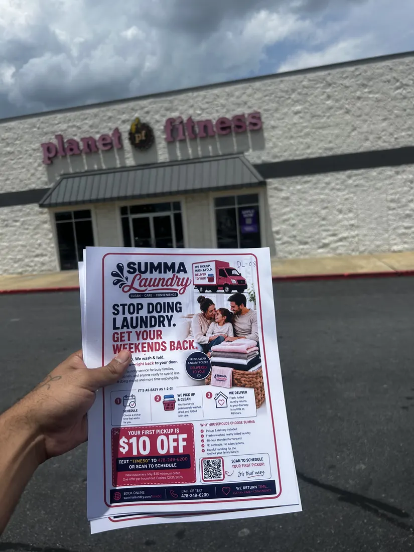 Una mano sosteniendo flyers impresos de Summa Laundry frente a un gimnasio Planet Fitness.